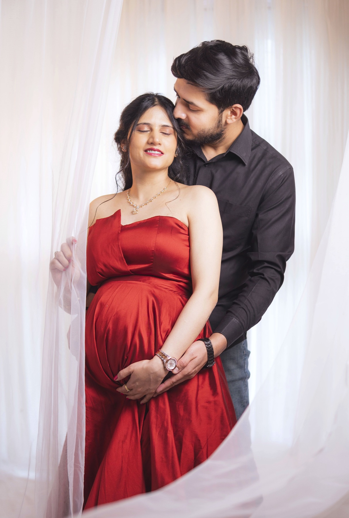 Indoor red gown maternity couple portrait with white drapes